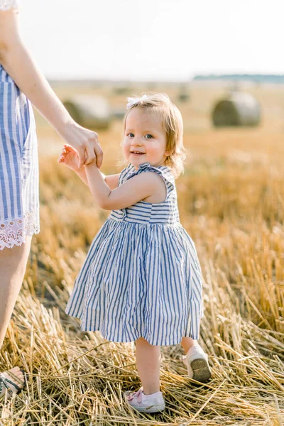 Retrato emocional de uma menina feliz e alegre em vestido listrado, segurando a mão de sua mãe enquanto caminhavam juntos em um campo de trigo com fardos de feno nos raios do pôr do sol. Verão. Infância — Fotografia de Stock