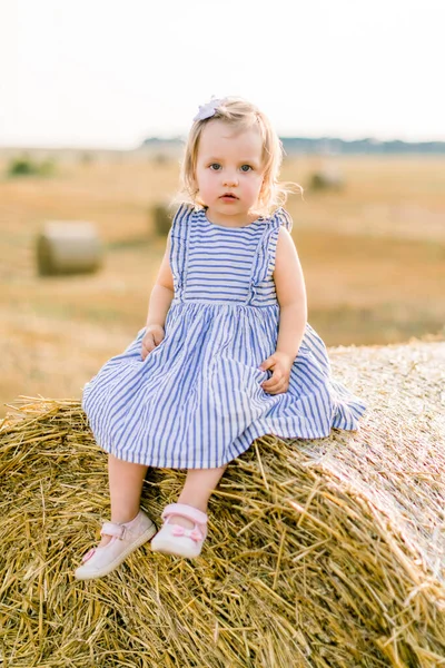 Κοντινό πλάνο πορτρέτο του χαριτωμένο όμορφο κοριτσάκι σε ριγέ φόρεμα κάθεται σε άχυρο στοίβα ή μπάλα σε κίτρινο φόντο τομέα σιτάρι την ηλιόλουστη μέρα του καλοκαιριού. Μωρό σε μπάλες σανού κατά τη διάρκεια της θερινής συγκομιδής — Φωτογραφία Αρχείου