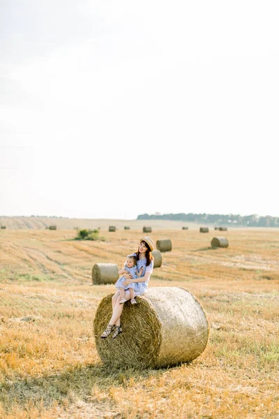 幸せな家族、母と小さな娘は、晴れた夏の日に黄色い小麦畑の干し草の山やベールに座っています。ママとかわいい赤ちゃん女の子でストライプドレスで楽しい夏の収穫時間 — ストック写真