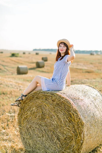 Menina de cabelos vermelhos encantador em chapéu e verão vestido listrado sentado em um fardo de feno no verão quente dia ensolarado, campo de trigo no fundo. Mulher olhando para a câmera e sorrindo, curtindo o verão — Fotografia de Stock