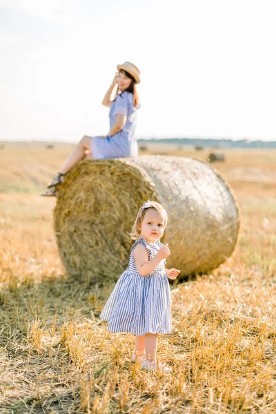 Sıcak ve güneşli yaz akşamlarında saman balyalarıyla buğday tarlasında küçük mutlu bir kız çocuğu. Elinde buğday dikenleri, elinde saman şapkası ve elbisesiyle güzel annesi arka planda saman yığınının üzerinde otururken. — Stok fotoğraf