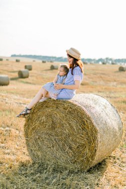 Çizgili elbiseli ve hasır şapkalı genç bir kadın saman balyasında oturur ve sevimli küçük kızına sarılır. Yaz günbatımının tadını samanlıklarla çıkarır.
