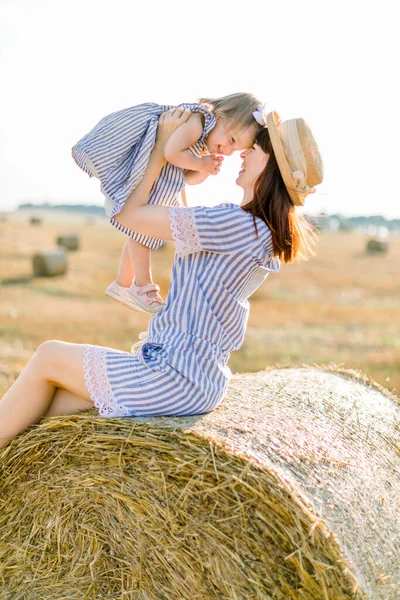 Κοντινό πλάνο του χαρούμενη μητέρα γέλιο και το κοριτσάκι, κάθεται σε άχυρο στοίβα ή μπάλα σε κίτρινο σιτάρι τομέα την ηλιόλουστη μέρα του καλοκαιριού. Μαμά και κόρη παίζουν, κοιτάζονται, κατά τη διάρκεια του καλοκαιριού — Φωτογραφία Αρχείου