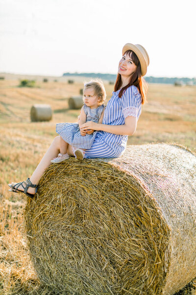 Young beautiful woman in striped dress and straw hat sits on hay bale and hugs her adorable little baby girl, enjoying summer sunset in field with haystacks