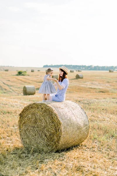 Criança feliz menina de dois anos de idade em vestido listrado brincando com a jovem mãe sorridente no campo de outono de verão com fardos de feno, sentados juntos na pilha de feno. Retrato do pôr do sol de verão — Fotografia de Stock