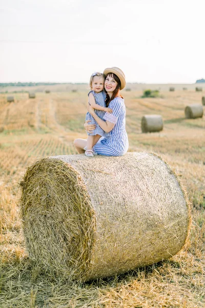 Mavi çizgili elbiseli mutlu küçük kız çocuğu yaz buğday tarlasında samanlıkta saman balyasında oturan genç annesine sarılıyor. — Stok fotoğraf