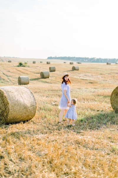 Summer bright portrait of little baby girl playing with her mom, wearing similar striped dresses, in beautiful field with hay bales on the background. summer, sunny walking, happy childhood — Stock Photo, Image