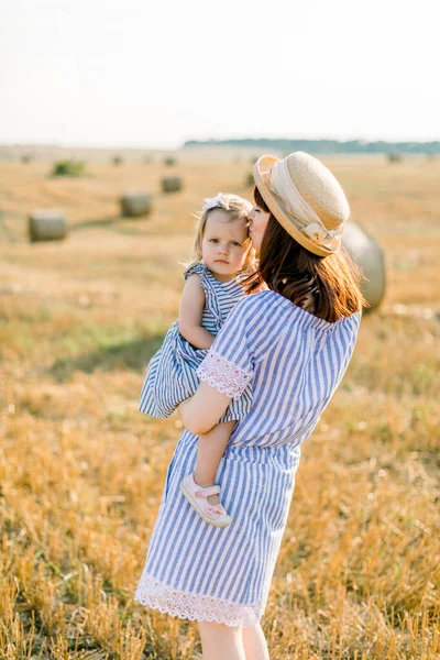 Güzel, beyaz, sevimli, küçük kızını elinde tutan, mavi çizgili elbiseler giyen, açık sarı buğday tarlasında bir kamış ve saman balyasıyla yürüyen bir anne. — Stok fotoğraf