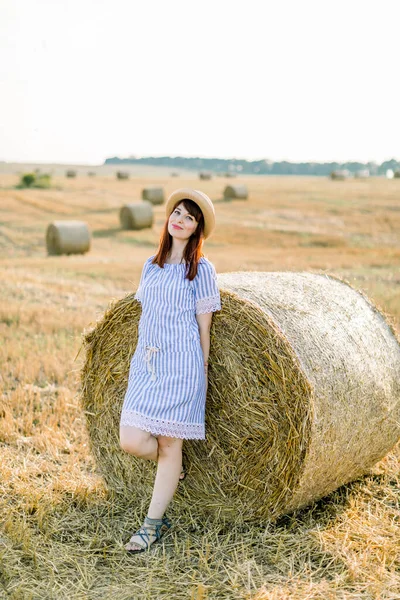 Paisagem rural do campo de trigo agrícola ao pôr-do-sol de verão. Mulher branca feliz muito jovem em chapéu de palha e vestido listrado azul posando perto do fardo de feno e desfrutando de noite quente e férias — Fotografia de Stock