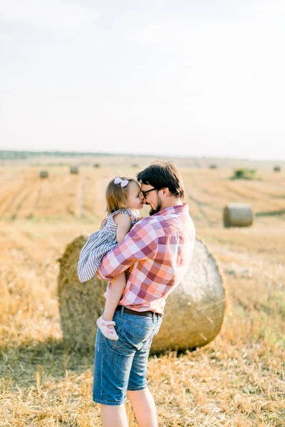 Joyeux jeune père joyeux, bel homme barbu en jeans et chemise à carreaux, tenant sur les mains sa jolie petite fille en robe, embrassant et touchant les fronts, marchant dans le champ à côté de balles de foin — Photo