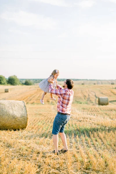 若い髭を生やした父親がかわいい娘と一緒に踊り、手をつないで夏の夕日のフィールドで干し草の玉を背景に楽しんでいます。屋外夏の家族のショット — ストック写真