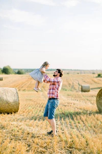 Yakışıklı, genç, sakallı bir baba, küçük tatlı kızıyla dans ediyor, onu elinde tutuyor, yaz günbatımı tarlasında arka planda saman balyalarıyla eğleniyor. Açık havada yaz ailesi vuruldu. — Stok fotoğraf