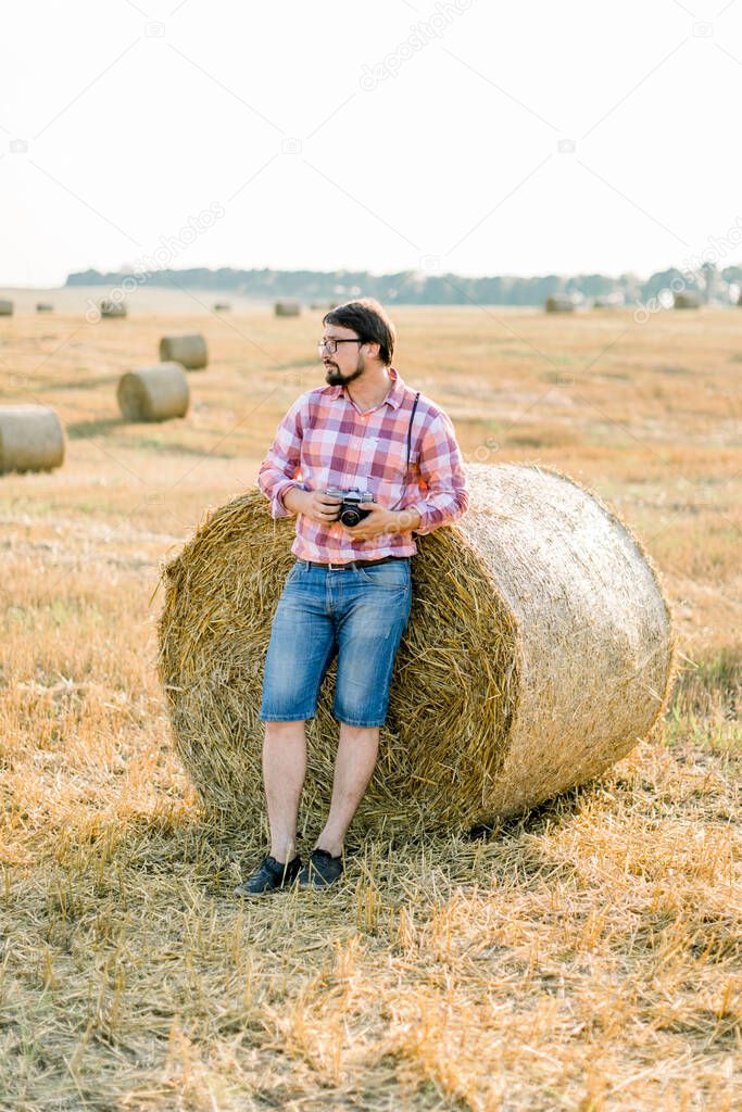 Handsome bearded brunette man, wearing checkered shirt and jeans leaning on a hay bale in goldern wheat field, posing, holding retro camera in hands, and enjoying warm sunset in summer day