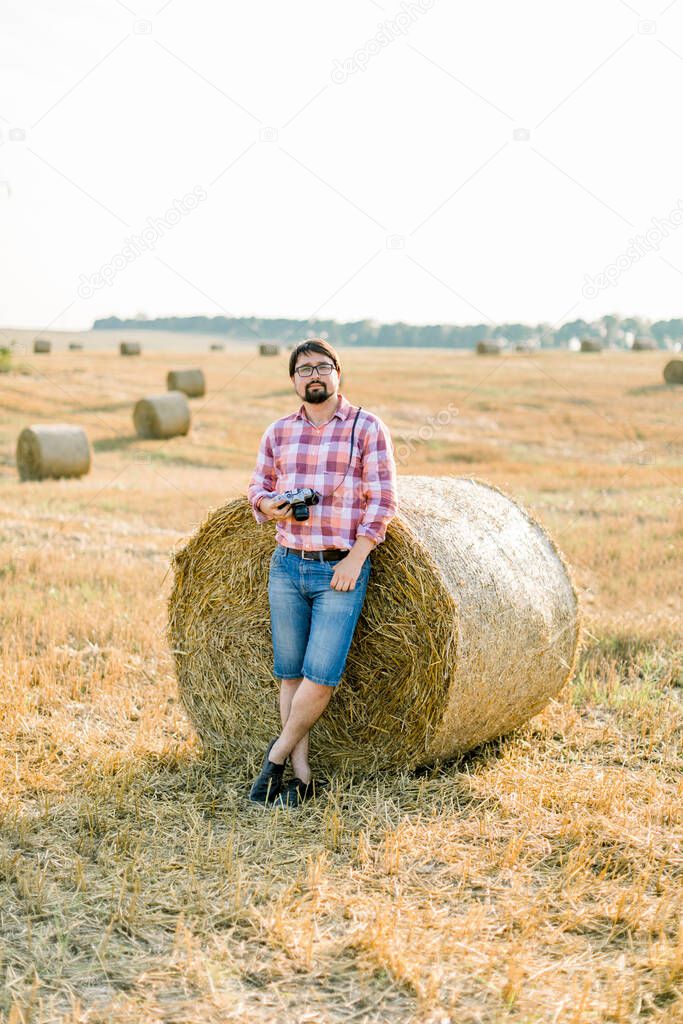 Handsome bearded brunette man, wearing checkered shirt and jeans leaning on a hay bale in goldern wheat field, posing, holding retro camera in hands, and enjoying warm sunset in summer day