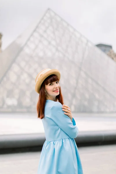 PARIS, FRANÇA - 17 de setembro de 2019: Uma jovem e elegante ruiva de vestido azul, posando para a câmera em frente à pirâmide de vidro no Museu do Louvre — Fotografia de Stock