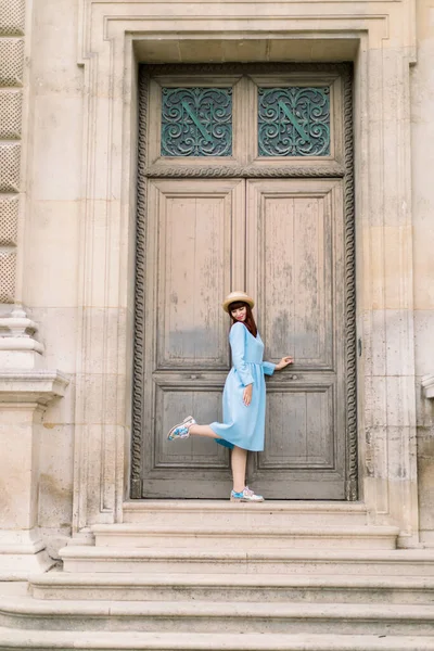 Charming elegant young woman wearing blue dress, posing in front of ancient wooden door of old vintage building with one leg up