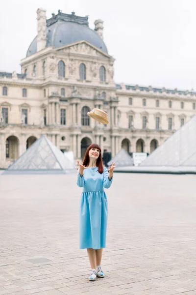 PARIGI, FRANCIA - 17 settembre 2019: Giovane donna in abito blu che vomita il cappello, in visita al museo del Louvre, in piedi di fronte alle famose piramidi di vetro . — Foto Stock