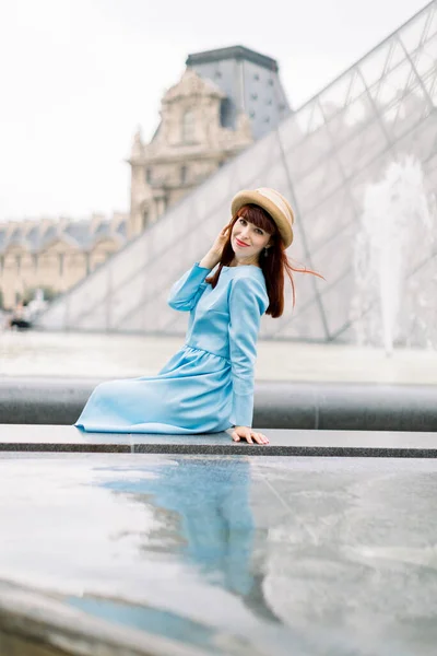 PARIS, FRANÇA - 17 de setembro de 2019: Jovem mulher feliz de vestido azul, sentada perto da fonte e posando para uma foto com o fundo da pirâmide de vidro no museu do Louvre — Fotografia de Stock