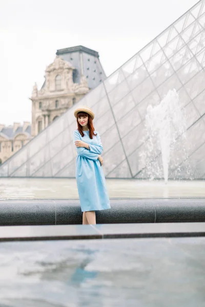 PARIGI, FRANCIA - 17 settembre 2019: Giovane donna felice in abito blu, posa vicino alla fontana e guarda la macchina fotografica, con lo sfondo piramidale di vetro al Museo del Louvre — Foto Stock