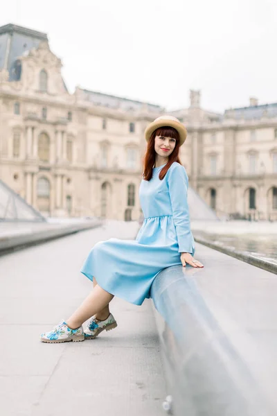 PARIGI, FRANCIA - 17 settembre 2019: Giovane donna felice in abito blu, seduta vicino alla fontana e in posa per una foto con lo sfondo della piramide di vetro al Museo del Louvre — Foto Stock