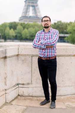 Gözlüklü, sakallı, sıradan kıyafetler ve pantolonlar giyen, kameraya bir fotoğraf için poz veren, Eiffel Kulesi 'nin önündeki Seine Nehri köprüsünde duran yakışıklı esmer adam.