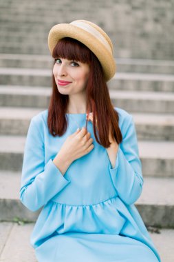 Mavi elbiseli ve şapkalı güzel kızıl saçlı kadın şehir merdivenlerinde otururken kameraya poz veriyor. Eski Avrupa şehrinde merdivenlerde bir kadın