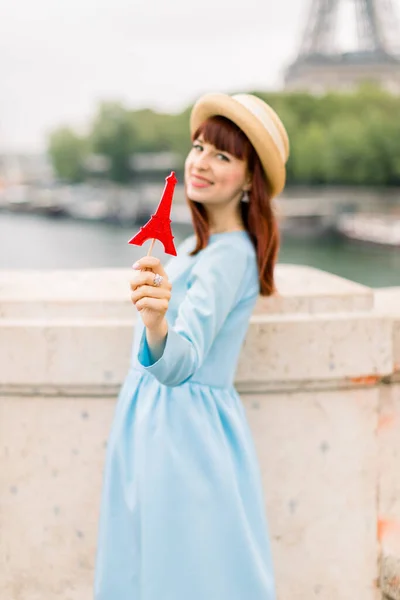 Módní portrét krásné rudovlasé dívky v slaměném klobouku a modrých šatech, ukazující krásné červené lízátko ve tvaru Eiffelovy věže a pozadí řeky Seiny. Soustřeď se na lízátko — Stock fotografie