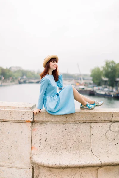 Mulher elegante jovem, usando chapéu de palha e vestido azul elegante, posando para câmera, enquanto desfruta de uma excelente vista sobre a torre Eiffel e Sena, sentado na ponte em Paris — Fotografia de Stock