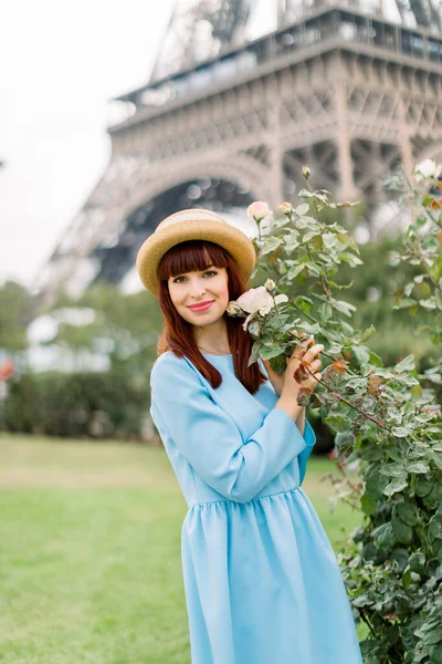 Bella giovane donna dai capelli rossi, indossa un abito elegante blu e un elegante cappello di paglia, in posa vicino ai cespugli di rose in fiore sulla vecchia città europea di Parigi all'aperto. Torre Eiffel sullo sfondo — Foto Stock