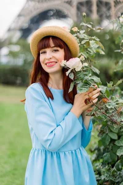 Portrait en gros plan d'une charmante jeune femme aux cheveux rouges souriante, portant une robe élégante bleue et un chapeau de paille élégant, posant près des rosiers à fleurs à Paris en plein air avec la tour Eiffel en arrière-plan — Photo