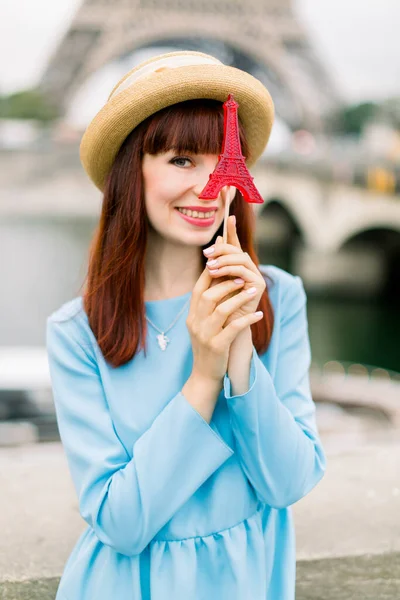 Mladá žena stojící na pařížské ulici před řekou Seinou, mostem a Eiffelovou věží, skrývající jedno oko s červeným lízátkem, usmívající se do kamery — Stock fotografie