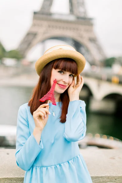 Bella ragazza con rossetto rosso con cappello di paglia e vestito blu, tiene lecca-lecca rosso, sorride alla macchina fotografica, in piedi all'aperto sullo sfondo del fiume Senna, ponte e torre Eiffel — Foto Stock