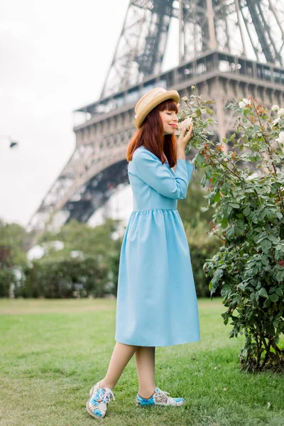 Jovem mulher bonita, vestida com elegante vestido azul claro e chapéu de palha, caminha na rua da cidade de Paris, perto dos arbustos floridos rosa, tocando e cheirando as flores. Torre Eiffel em segundo plano — Fotografia de Stock