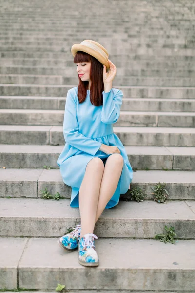 Krásná mladá zrzavá žena v modrých šatech a klobouku, pózující na kameru, zatímco sedí venku na městských schodech. Žena on stairs in old European city — Stock fotografie