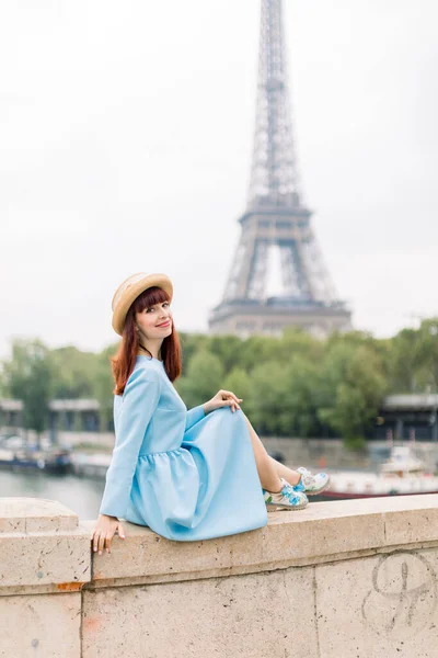 Mulher elegante jovem, usando chapéu de palha e vestido azul elegante, posando para câmera, enquanto desfruta de uma excelente vista sobre a torre Eiffel e Sena, sentado na ponte em Paris — Fotografia de Stock