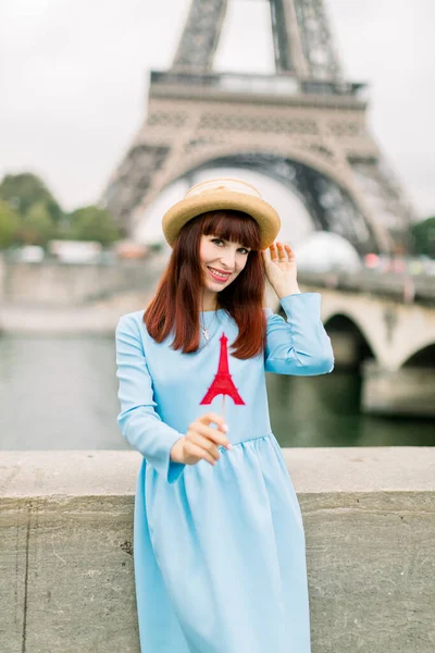 Mladá atraktivní usměvavá žena v slamáku a modrých šatech, s lízátkem ve tvaru Eiffelovy věže, stojící před Eiffelovou věží a řekou Seinou. Cestovní koncept — Stock fotografie