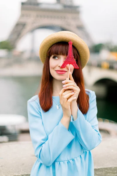 Beautiful woman with sweet candy lollipop posing on the background of Eiffel tower and Seine river. Outdoor photo of happy red-haired girl with lollipop looking aside and hiding her eye with candy
