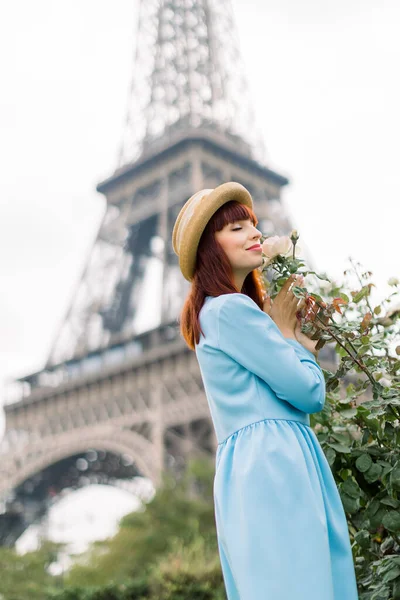 Romantická fotka krásné mladé Francouzky se zavřenýma očima, jak si užívá vůni růžového květu, když kráčí venku v Paříži, stojí před Eiffelovou věží. Červené vlasy dívka v klobouku v blízkosti růží — Stock fotografie