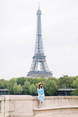 Mavi elbiseli genç kadın Paris ve Seine nehrinin manzarasının keyfini çıkarırken Eyfel kulesinin arka planındaki köprüde oturuyor.