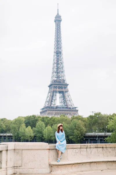 Junge Frau in blauem Kleid genießt die schöne Aussicht auf die Landschaft von Paris und die Seine, während sie auf der Brücke vor dem Eiffelturm sitzt — Stockfoto