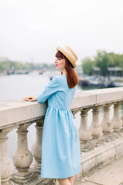 Jovem mulher atraente em vestido azul e chapéu de palha, desfrutando de ótima vista sobre o rio Paris e Sena, de pé na ponte e olhando para longe — Fotografia de Stock