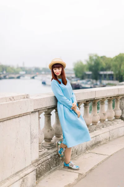 Giovane donna caucasica sorridente in abito blu e cappello di paglia, in piedi sul ponte e appoggiata sulla ringhiera, godendo di una splendida vista sul fiume Parigi e la Senna — Foto Stock