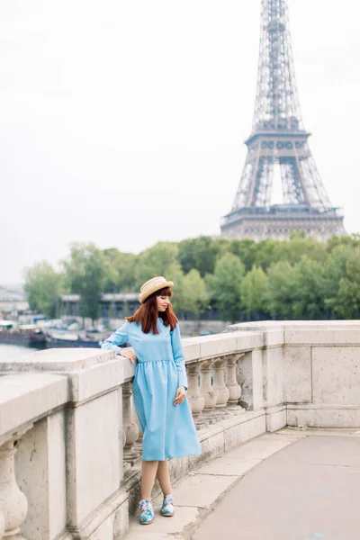 Okouzlující elegantní žena v modrých šatech, opírající se o most v Paříži ve Francii. Eiffelova věž a řeka Seina v pozadí. — Stock fotografie