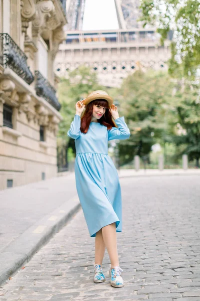 Vista di strada di bella giovane signora dai capelli rossi, con elegante abito blu e cappello di paglia, camminare durante la mattina a Parigi, in posa per la fotocamera. Torre Eiffel dietro la donna — Foto Stock