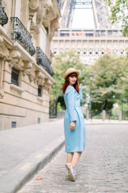 Mavi elbiseli ve şapkalı genç ve şık kızıl saçlı kadının arka planı. Arkasını dönüp kameraya bakıyor. Paris 'te karşıdan karşıya geçerken Eyfel Kulesi' ne doğru yürüyor.