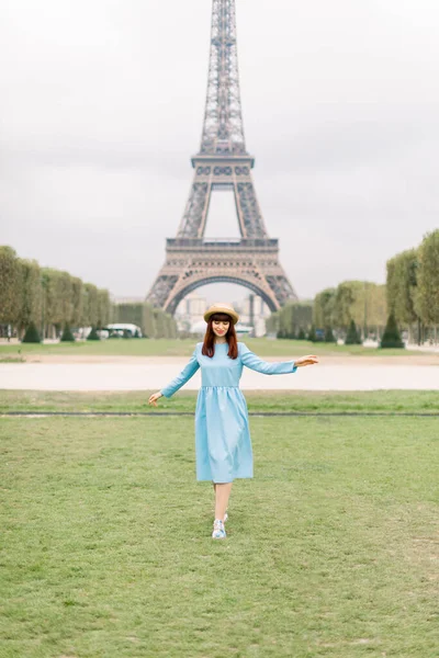 Menina europeia bonita vestindo vestido azul e chapéu de palha, desfrutando de férias em Paris, França no fundo Torre Eiffel. sorrindo e andando na grama verde no parque — Fotografia de Stock