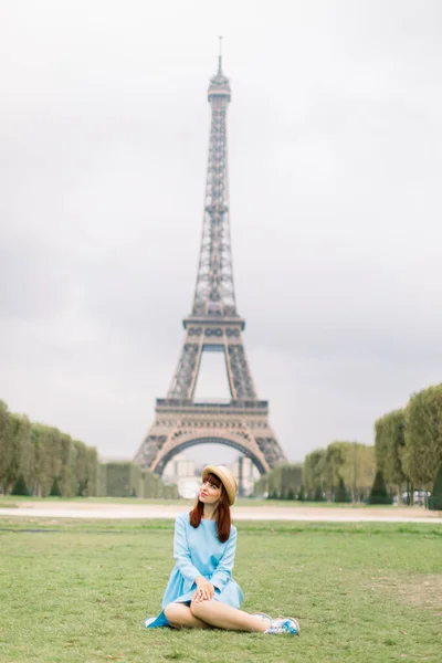 Elegante donna con cappello di paglia e vestito blu seduta sull'erba verde in un parco con la Torre Eiffel sullo sfondo — Foto Stock