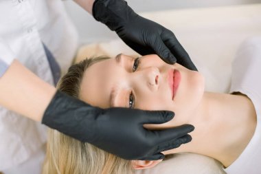 Güzellik salonunda kendini rahat hisseden gülümseyen genç kadına yüz masajı yapan kozmetik bilimci kadının elleri. Yaşlanma karşıtı yüz bakımı ve masaj kavramı