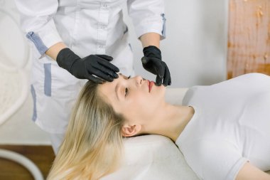 Kadın kozmetik uzmanının, güzellik salonunda kendini rahat hisseden gülümseyen genç kadına yüz masajı yaparken çekilmiş görüntüsü. Yaşlanma karşıtı yüz bakımı ve masaj kavramı