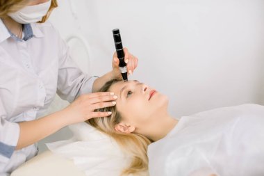 Modern dermatoloji kliniğinde kanepede yatan genç sarışın kız hasta cilt tanısı yaptırıyor. Dermatoskop kullanarak hastasının deri tipini inceleyen profesyonel bir kadın dermatolog.